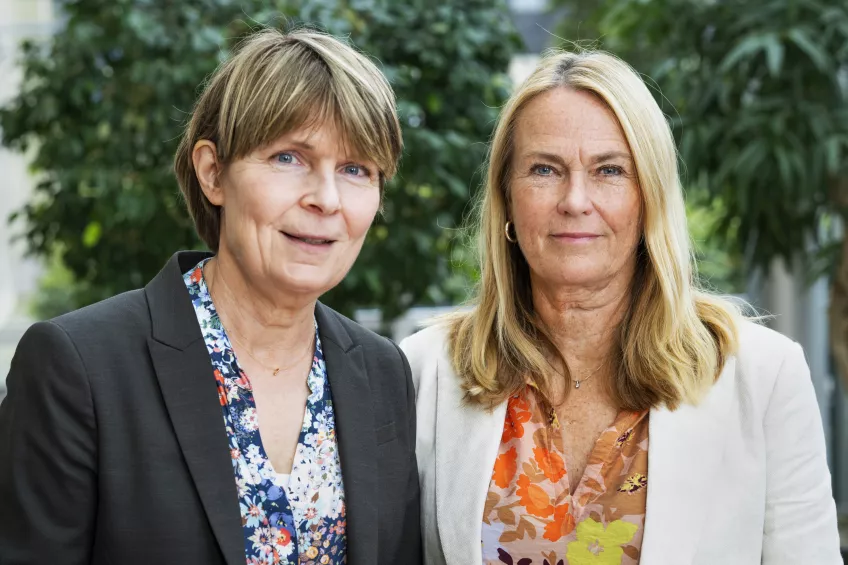 Två personer ståendes med grönska i bakgrunden. Foto.